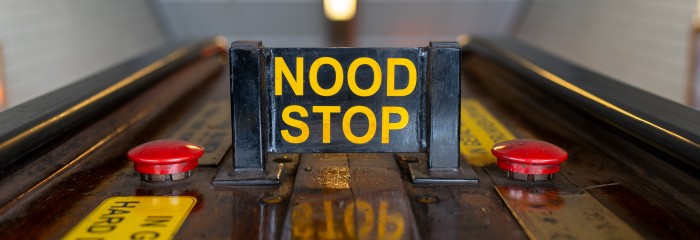 A sign on an escalator that reads "nood stop"  in yellow letters. "Nood stop" means "emergency stop" in Dutch. There are two red buttons on either side of the sign.
