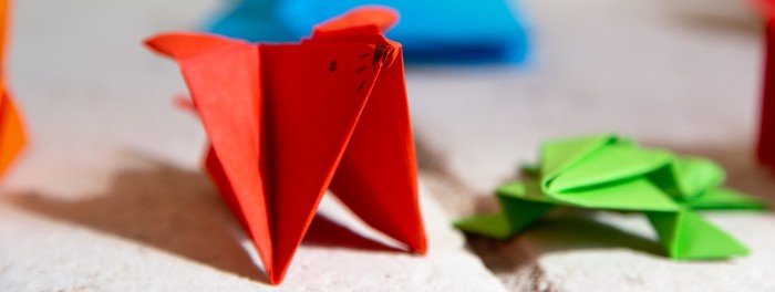 Colorful origami animals sitting on a stone bench. In the center of the frame is a small red mouse origami. To the right is a flat green frog origami.